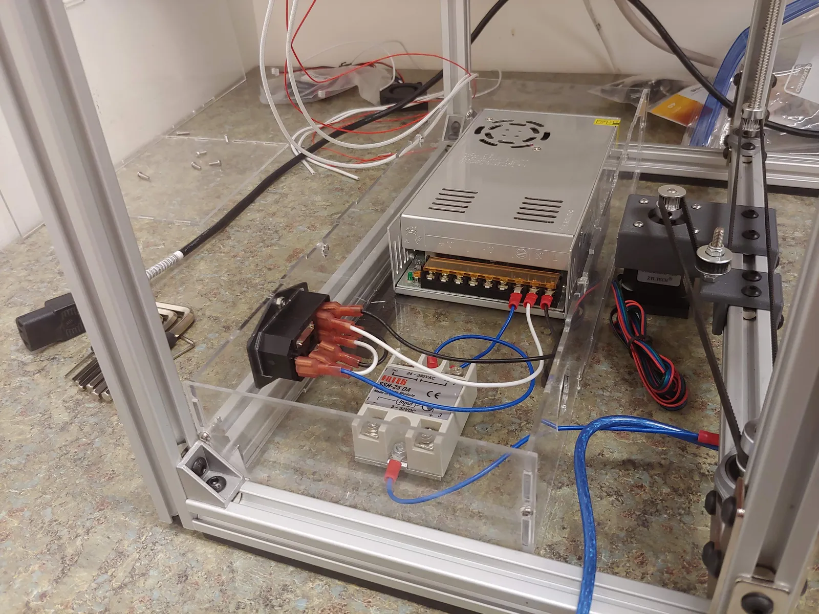 The rear electronics enclosure mounts the mains-voltage components, including the power input jack (with button and fuse), the 24V switching power supply, and the solid-state relay responsible for switching the AC-powered bed heater
